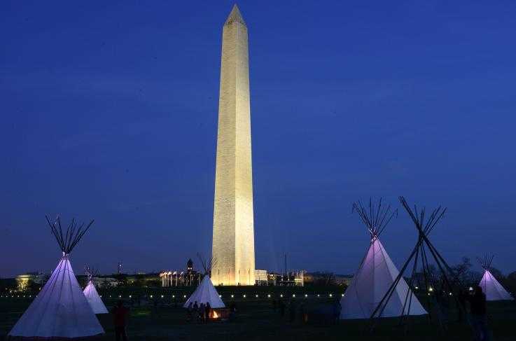 Four tipis and a campfire in the foreground on the National Mall at dusk with the Washington Monument, an obelisk, lit up in the background.