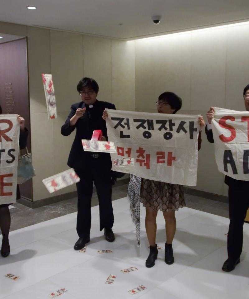Four people holding up signs saying 'Stop ADEX' in English and Korean. One of the people is wearing a dog collar.