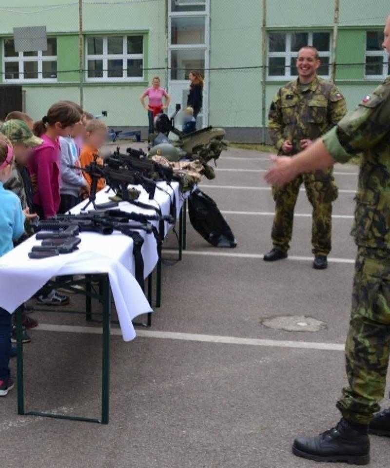 Soldiers talk to young children, who are stood in front of a table covered with guns.