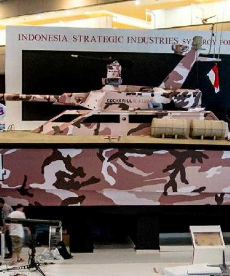 CMI Defences "tank boat" - a catamaran with a large turret, exhibited indoors at a trade show. The boat is painted in brown camoflage patterns.
