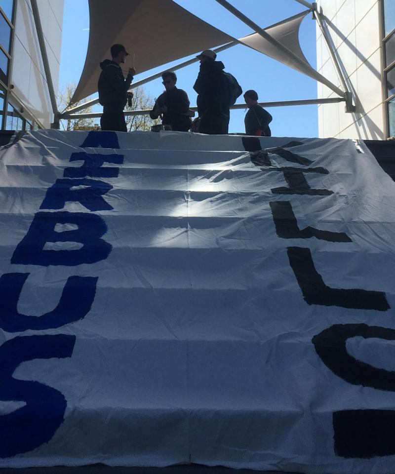 The main image is a set of steps outside, covered in a large banner reading "Airbus Kills". At the top of the steps stand four people sillouhetted against the sky. They are playing drums