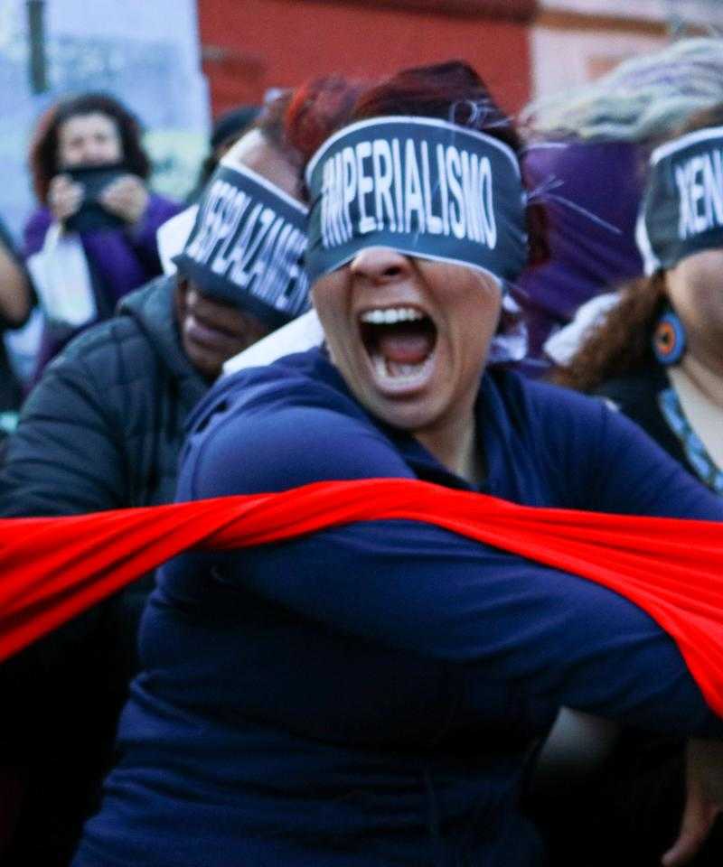 Una protesta. Una mujer en primer plano con una venda en los ojos que dice "imperialismo". Ella está tirando una tela roja. Detrás de ella hay otros manifestantes con los ojos vendados.