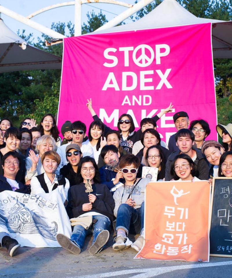 A group of korean activists gather in front of a pink sign saying "stop ADEX"