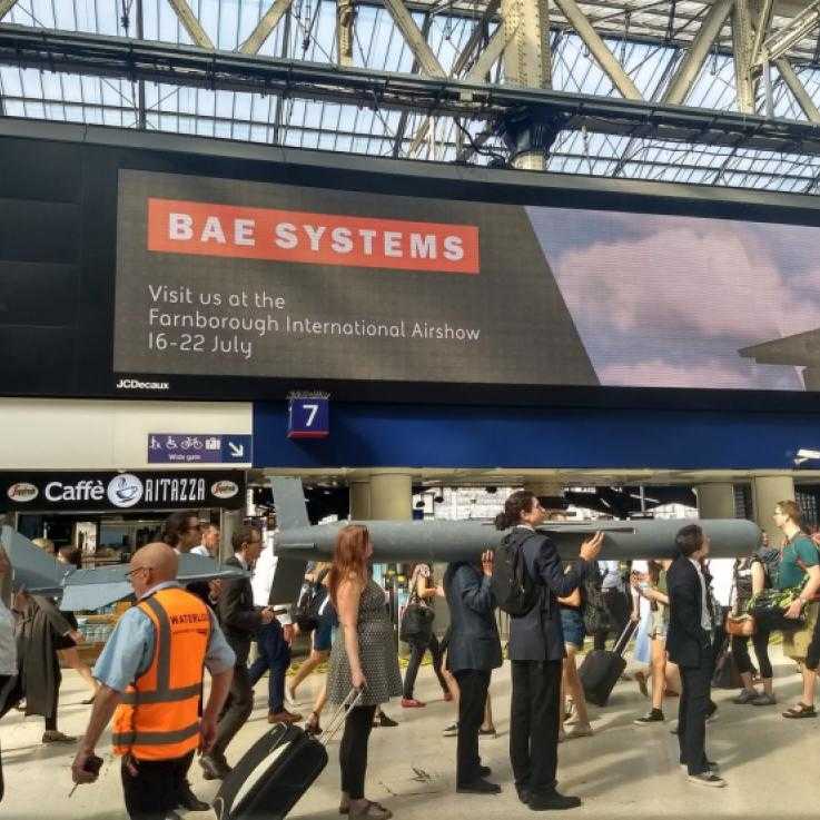 A train station lobby. A huge advertisement for BAE Systems is in the background. In the fore ground stands a line of activists dressed in suits, carrying a hgue missile on their shoulders.