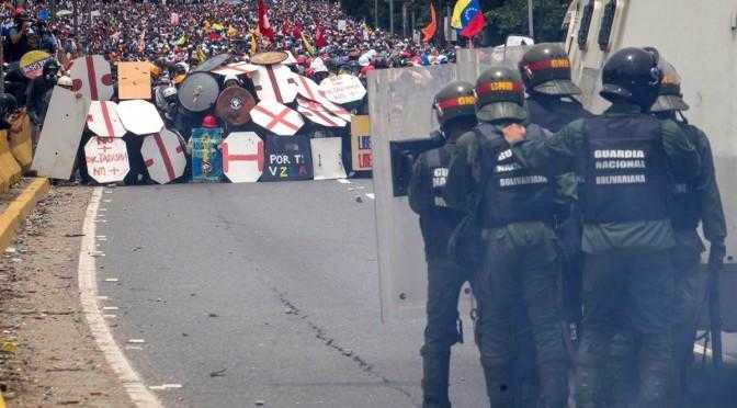 Police behind riot shields face a crowd of protesters behind a wall of homemade shields in Venezuela