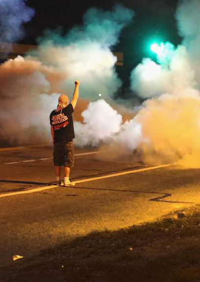 A man stands with his back to the camera and fist raised amidst clouds of tear gas