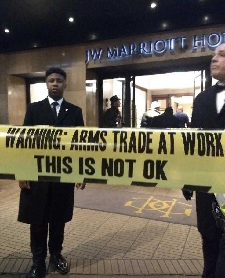 Security guards stand in front of a sign saying "arms trade at work this is not ok"