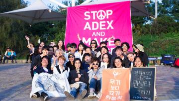 A group of korean activists gather in front of a pink sign saying "stop ADEX"