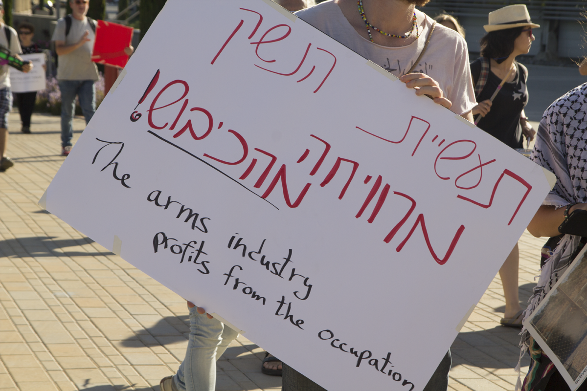 A protester outside the ISDEF arms fair holds a sign reading "The arms industry profits from the occupation"