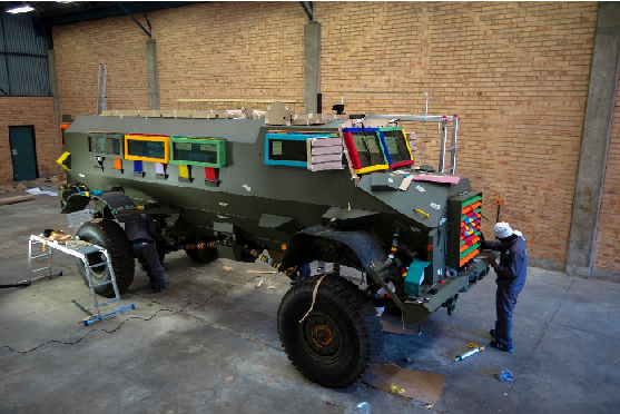 A largely undecorated Casspir stands in a warehouse with brick walls and a concrete floor.  The khaki paintwork of the Casspir is interrupted by bright, multicoloured beading on the window frames and grille.  An artist in overalls is working on the beading on the grille and near the back wheel there is a platform ladder.