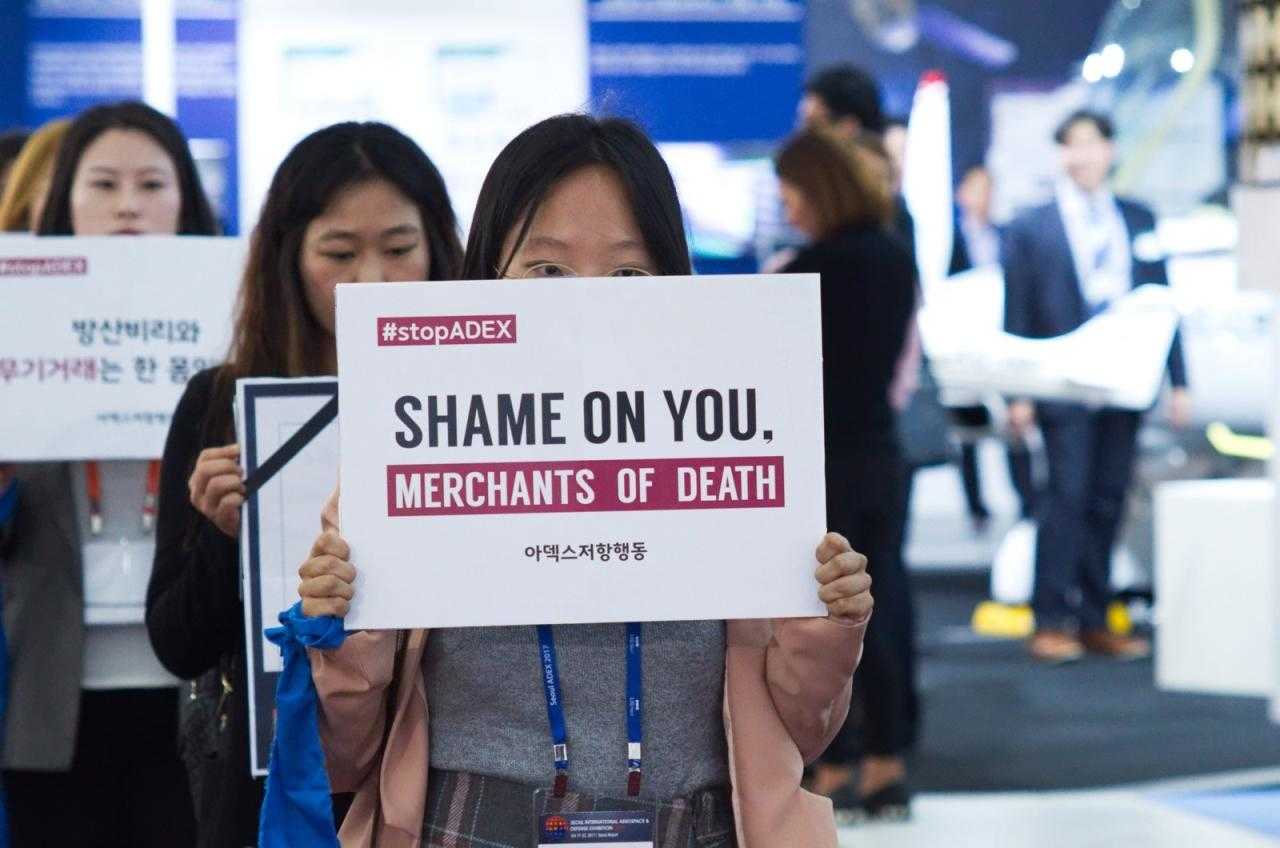 Mitglieder der Welt ohne Krieg ergreifen direkt auf einer Waffenmesse. Sie halten Schilder mit der Aufschrift "Schande über dich".