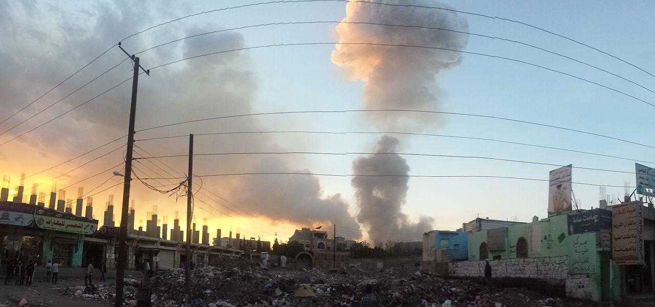 An airstrike in Sana'a, Yemen, in 2015. A cloud of smoke rises from behind buildings. The sun is rising to the left of the picture.