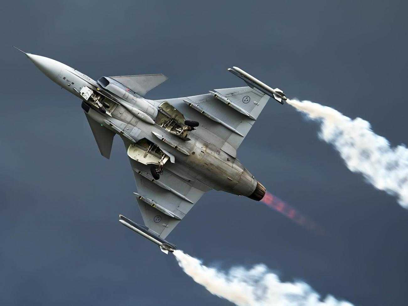 A fighter aircraft in the air during a display. The image is shot from below, and the aircraft has plumes of smoke trailing from the tip of each wing.