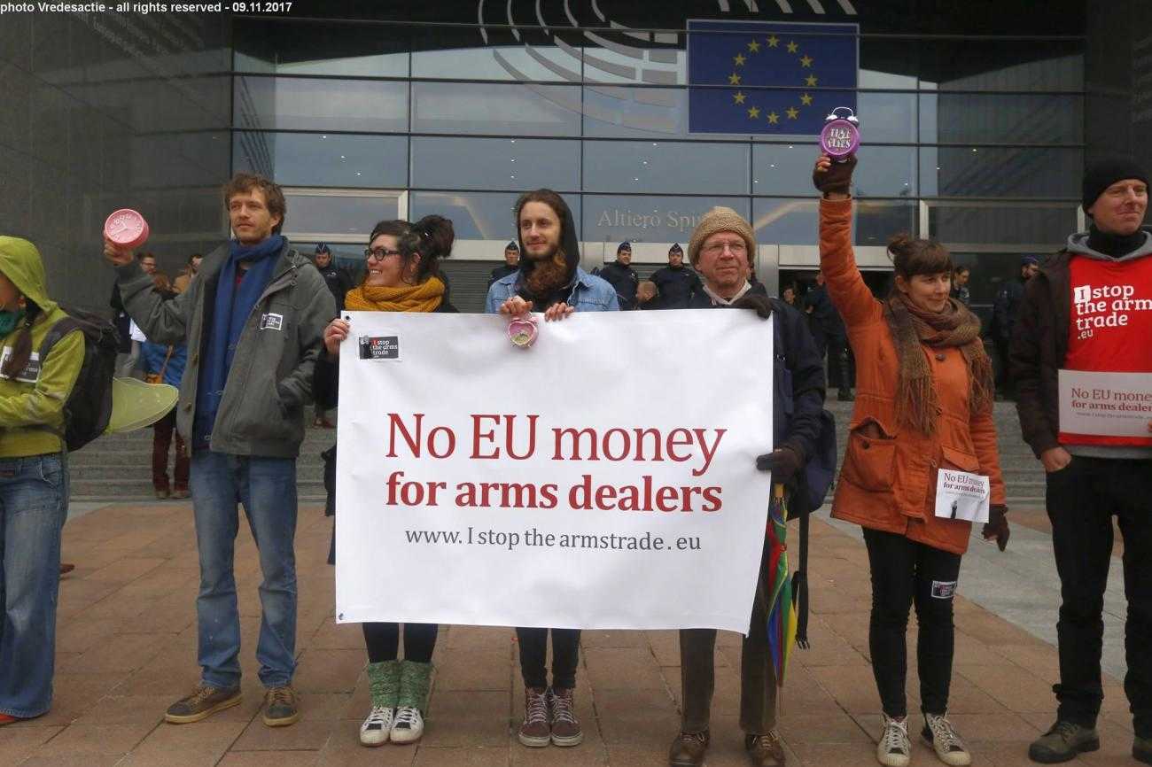 A line of campaigners stand with a banner reading "No EU money for arms dealers", in front of a building flying the EU flag