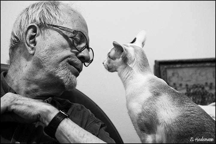 David McReynolds on the left side of the photo, wearing glasses, looking at a cat sat on the right.