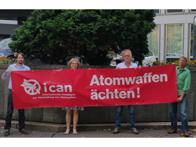 Four people stand with a red banner reading "Ban Nuclear Weapons" 