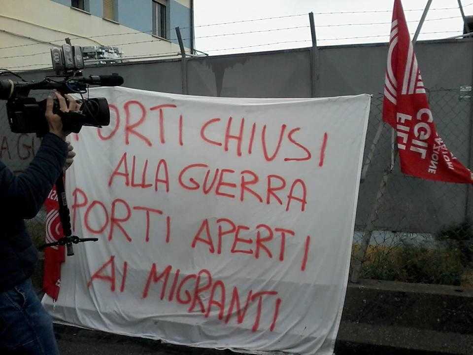 A banner reading "port closed to war, port open to migrants" in Italian, hanging off a fence