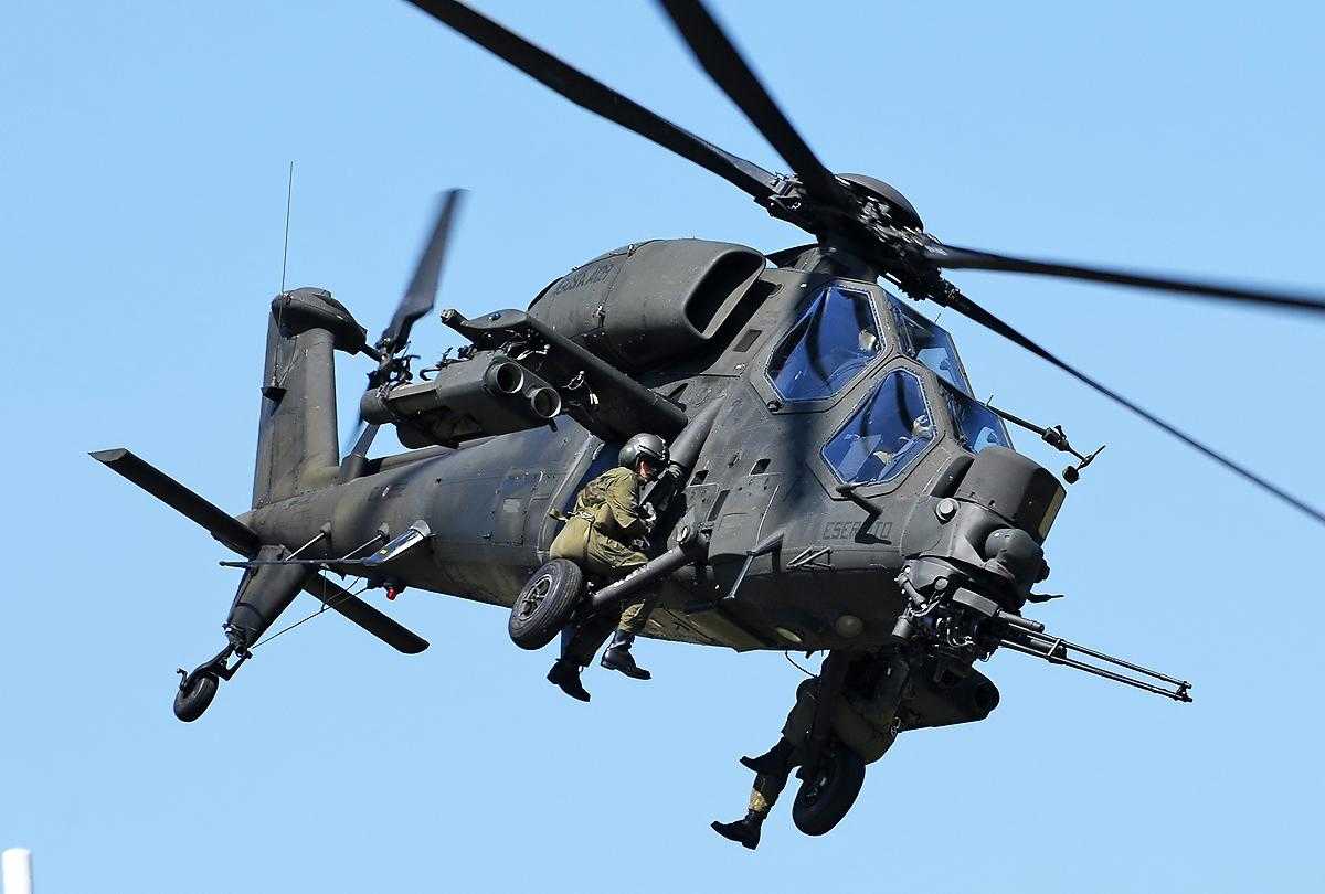 A military helicopter in flight. Two military personnel are sat on the outside.