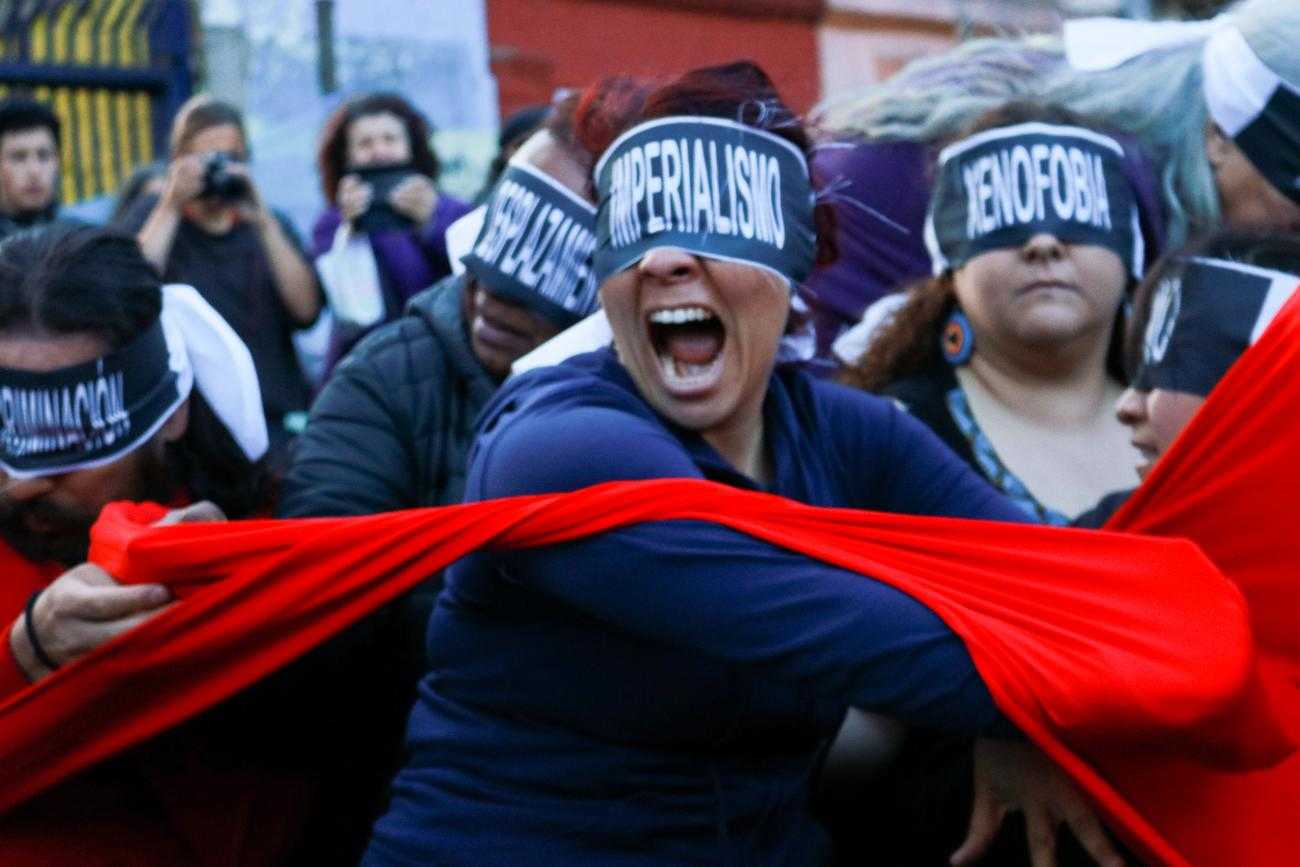 Una protesta. Una mujer en primer plano con una venda en los ojos que dice "imperialismo". Ella está tirando una tela roja. Detrás de ella hay otros manifestantes con los ojos vendados.