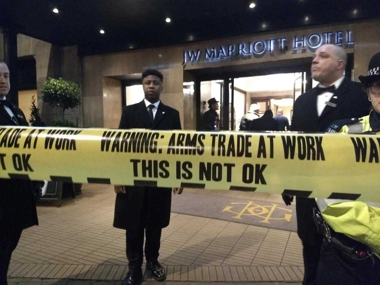 Three security guards stand at the door of a hotel. In the foreground is some fake security tape reading "Arms trade at work, this is not ok"