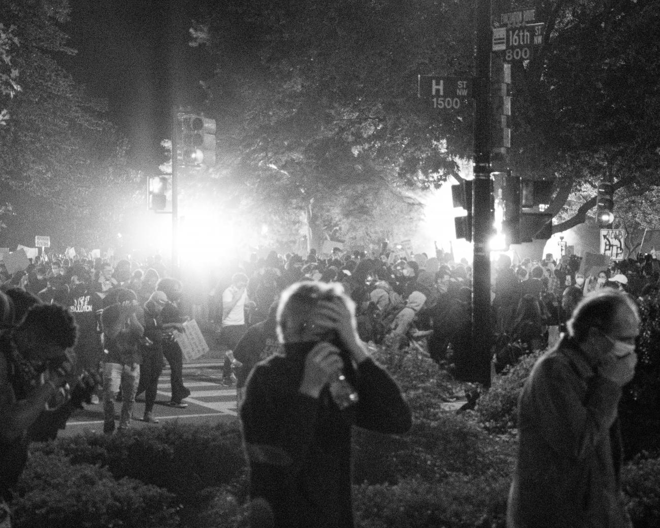 A black and white photo at night, showing large numbers of people walking through smoke with their mouths covered