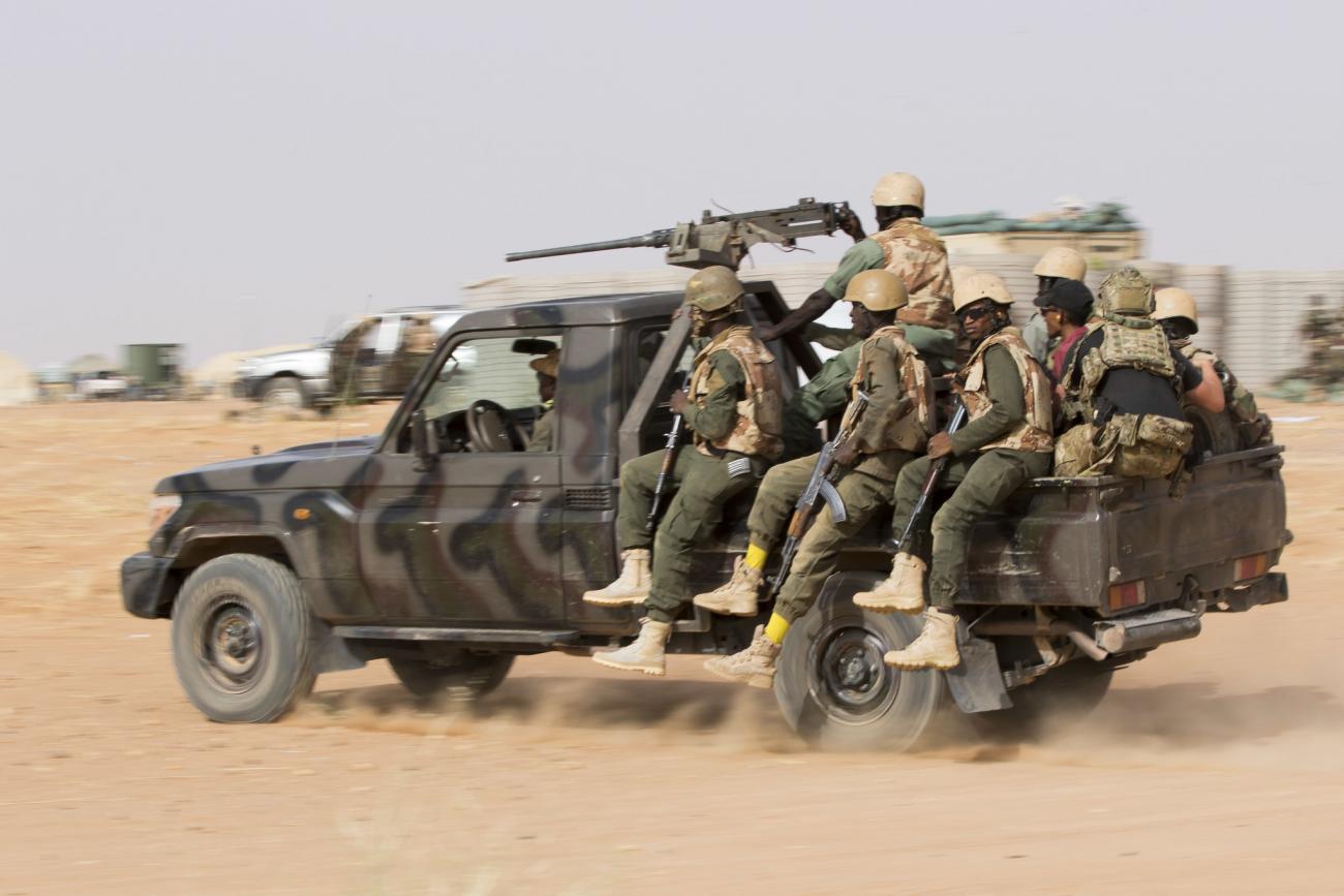A group of soldiers on a large jeep driving through the desert. The jeep has a large gun on top and the soldiers are armed.