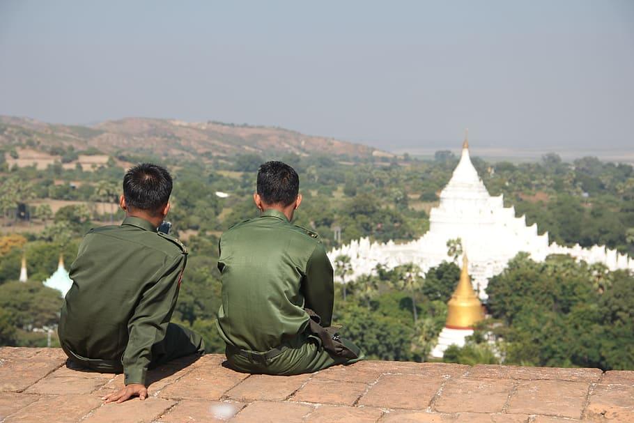 Two soldiers sitting with their backs turned, looking towards a river in the background.