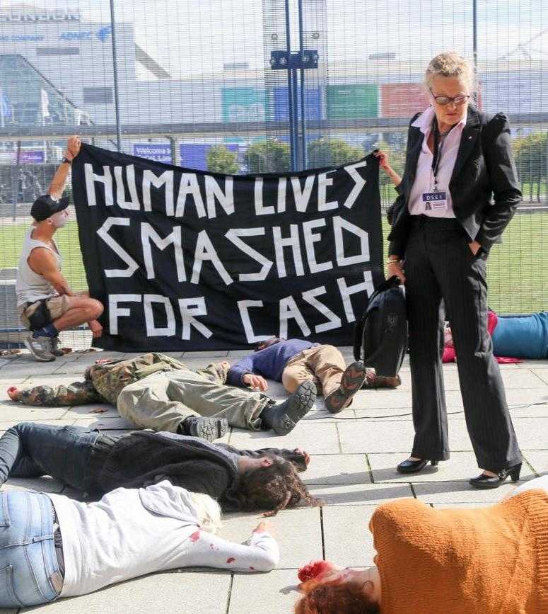 A die-in at DSEI 2017. People lie on the floor, while a woman dressed in business clothing looks down. Behind her activists hold a sign that reads "Human lives smashed for cash"