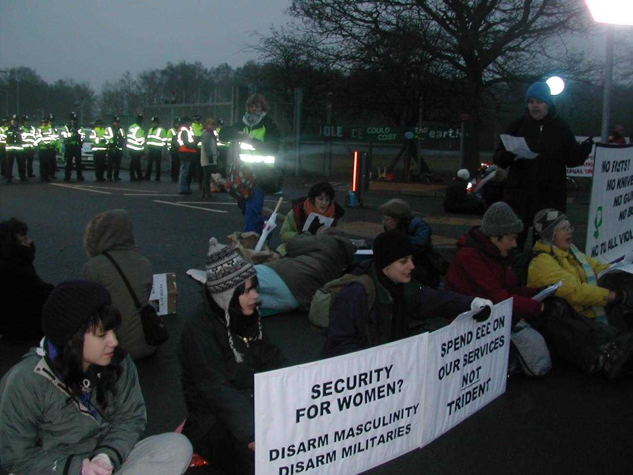 Women sit across a road in a protest against nuclear weapons. They are holding signs