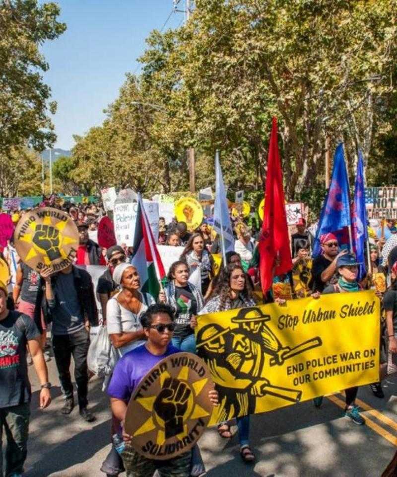 A crowd marches down a tree-lined avenue holding colourful banners and flags.  The yellow banner at the forefront of the picture reads 'Stop Urban Shield: End Police War on Our Communities'.