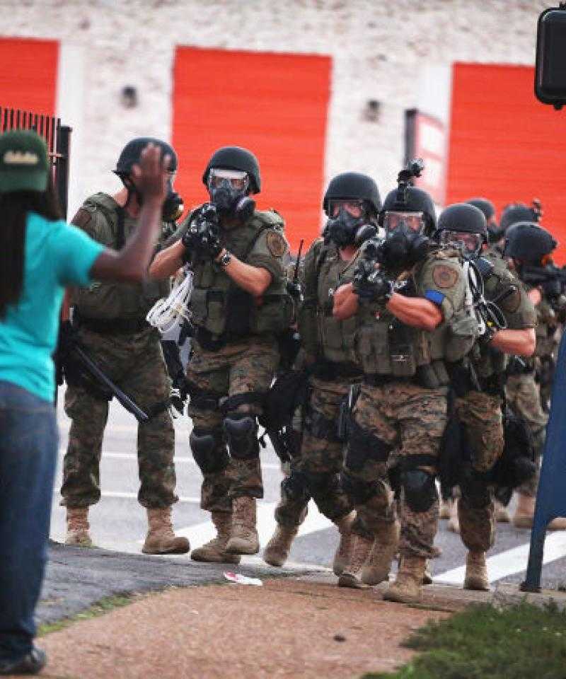 Heavily armed police pointing guns move towards a black person with their hands raised.  The back of a street sign reads 'Fuck the Police'.