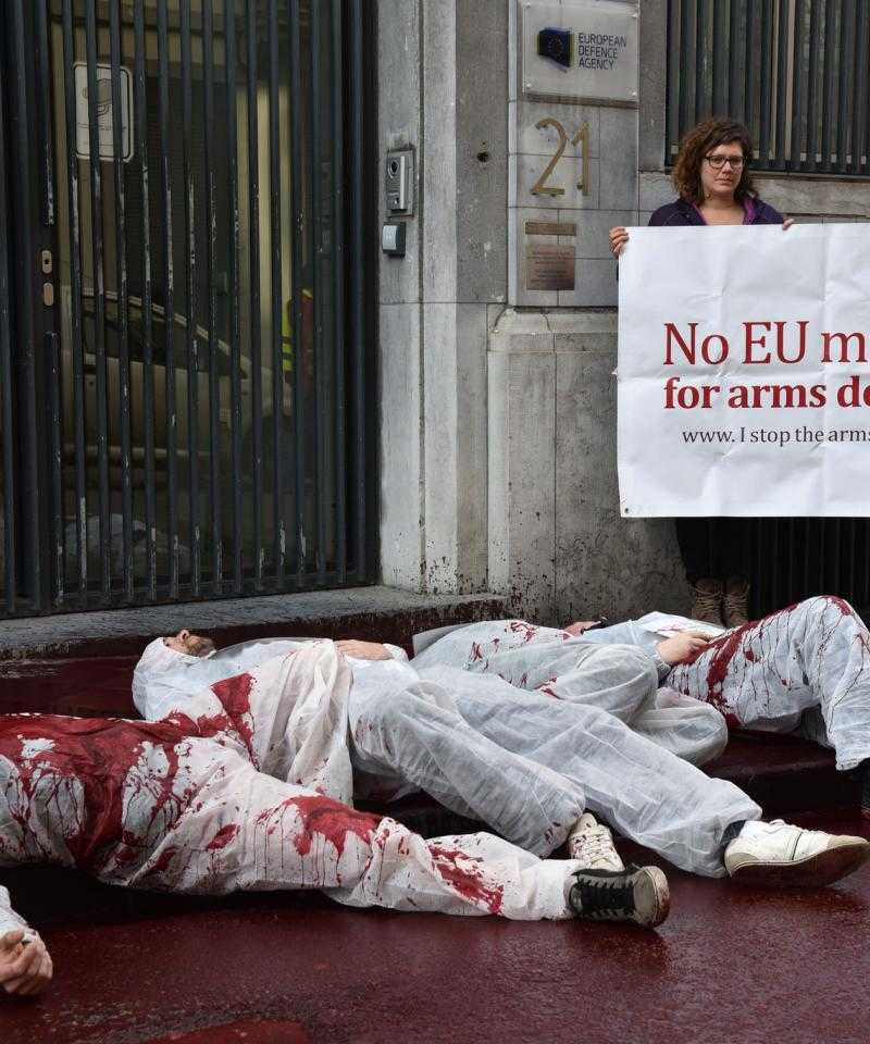 Three activists dressed in white overalls lie in a pool of fake blood. Behind them stand two more activists holding a banner reading "no money for arms dealers"
