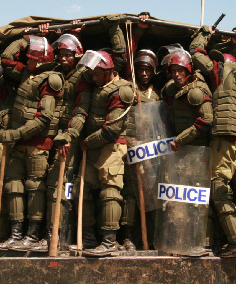 A large number of police armed with clubs wait to jump out of a van in Kenya. The police are wearing a lot of armour, shields, helmets and clubs.