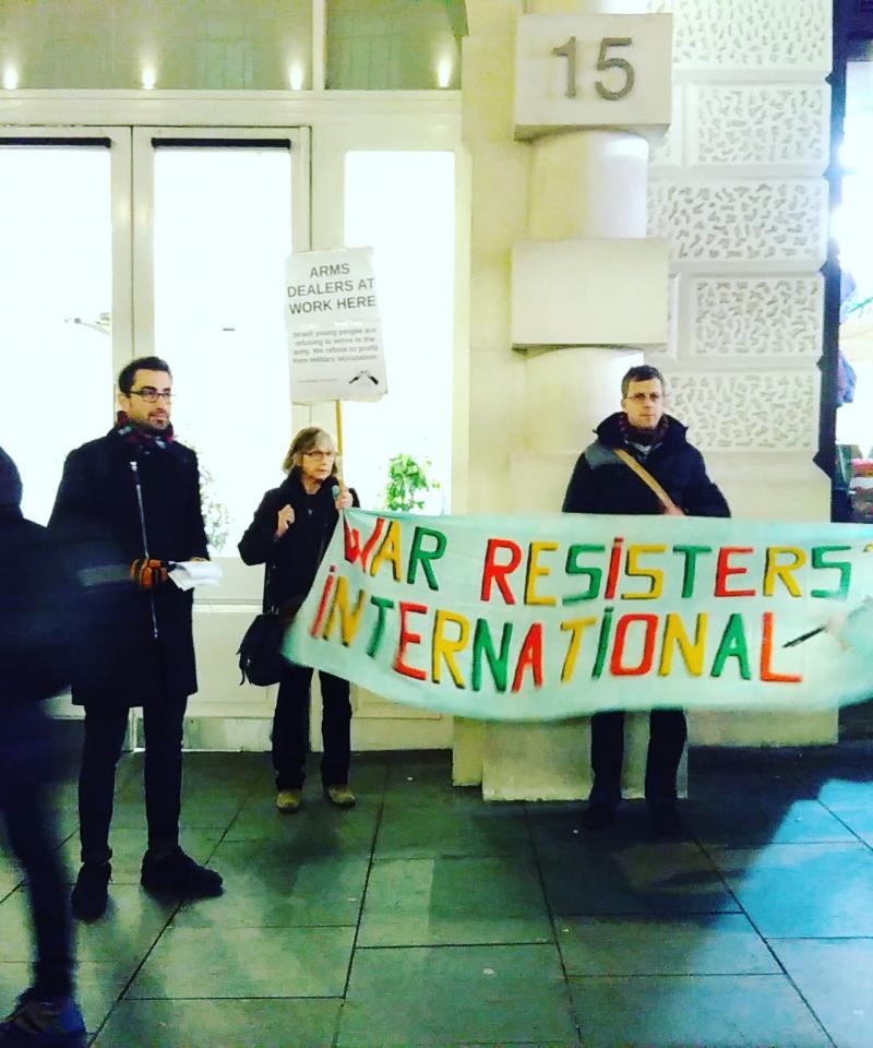 Several people holding a 'War Resisters' International' banner, and handing out leaflets, outside a building
