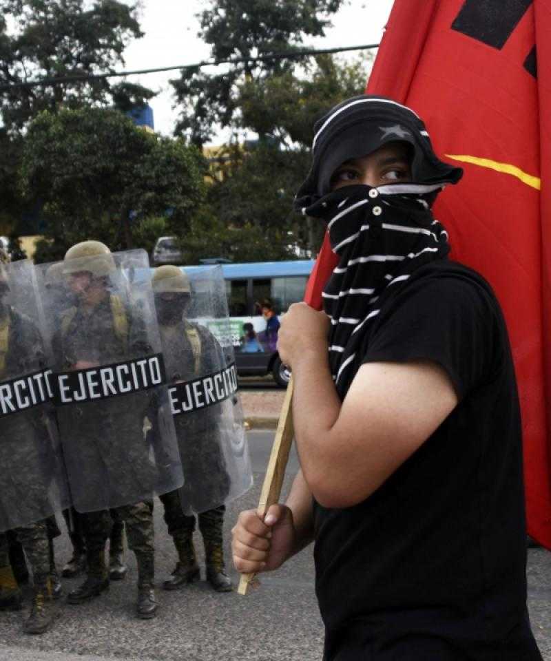 An activist with his face covered holds a red flag. He is stood in front of a line of police officers and military personnel holding shields.