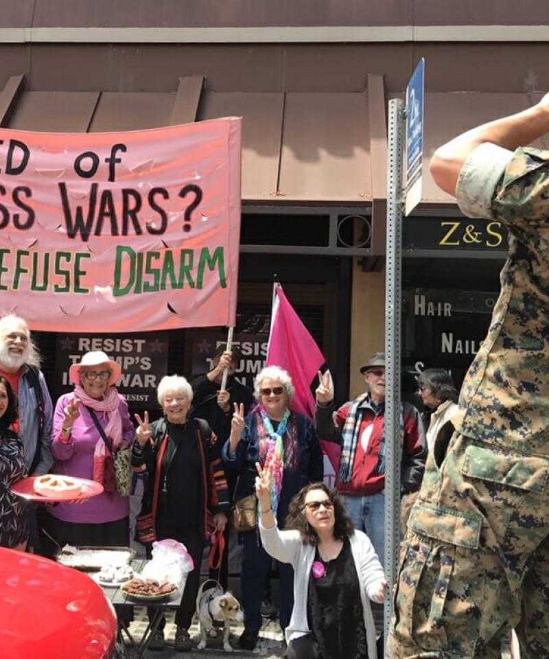 A group of people holding a pink banner saying 'Tired of Endless Wars? Resist, Refuse, Disarm' outside a recruitment centre, with a soldier taking a photo of them with a phone