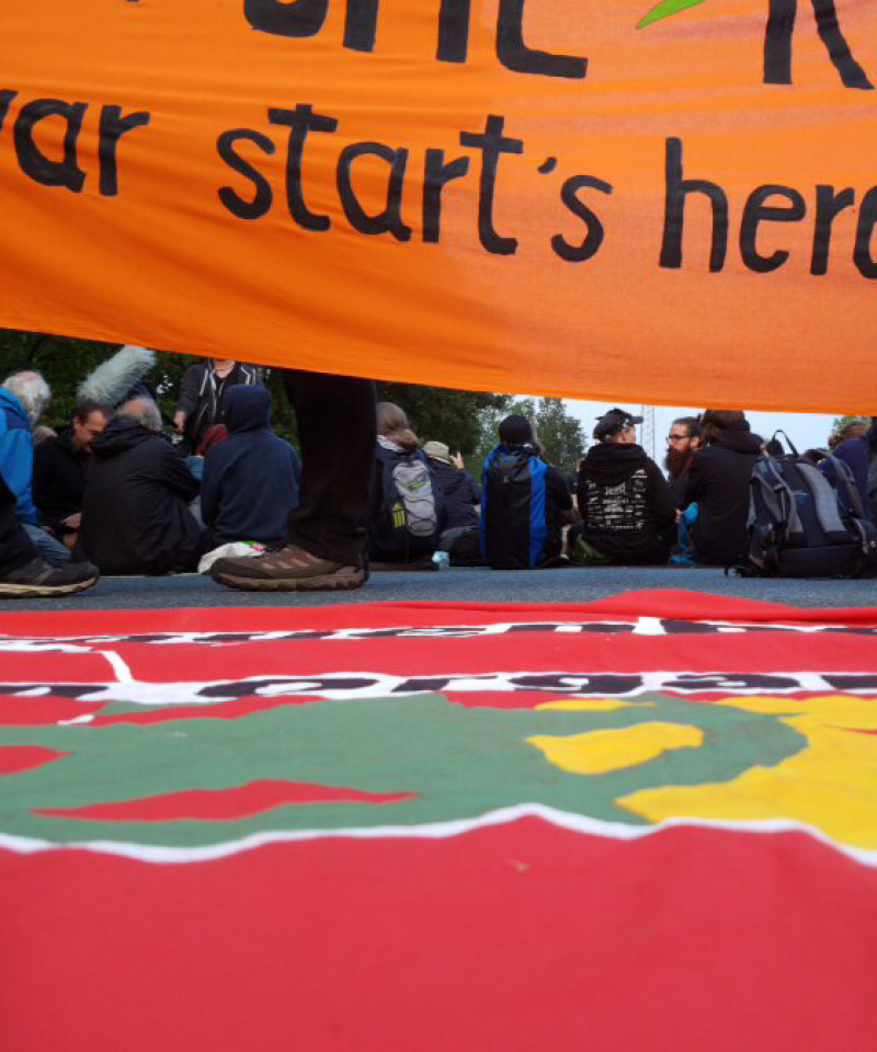 An orange banner at the top reads "War Starts Here". Another banner lies on the floor. Between them is a line of people sat with their backs to the camera, blocking a road.