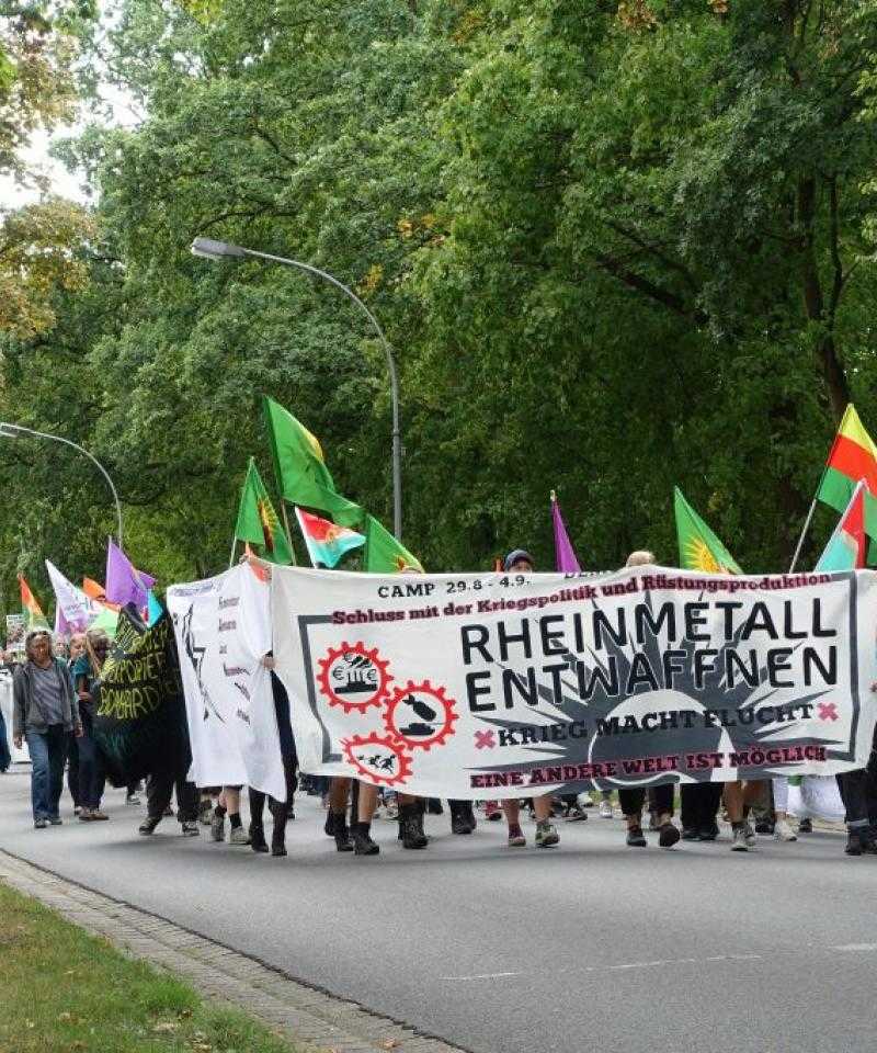 A large group of people march down a street. The front group are holding a banner reading "disarm Rheinmetall" in German