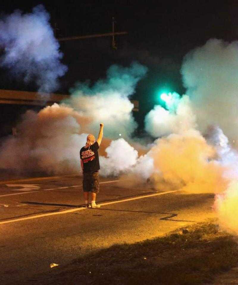 A man stands still with his arm raised amid a cloud of tear gas at night