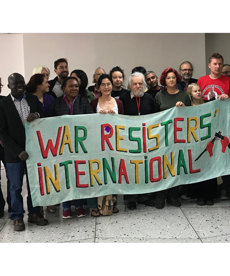 Two dozen people stand behind a banner reading "War Resisters' International"
