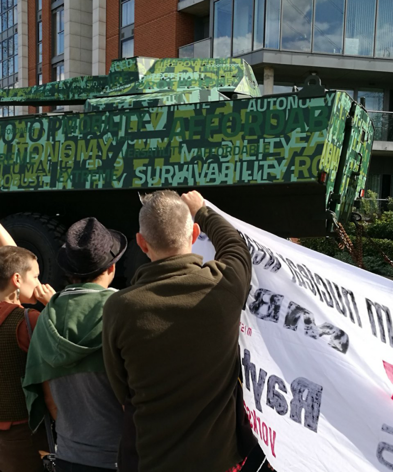 A tank entering the DSEI arms fair, covered in marketing slogans. In front stand a crowd holding banners