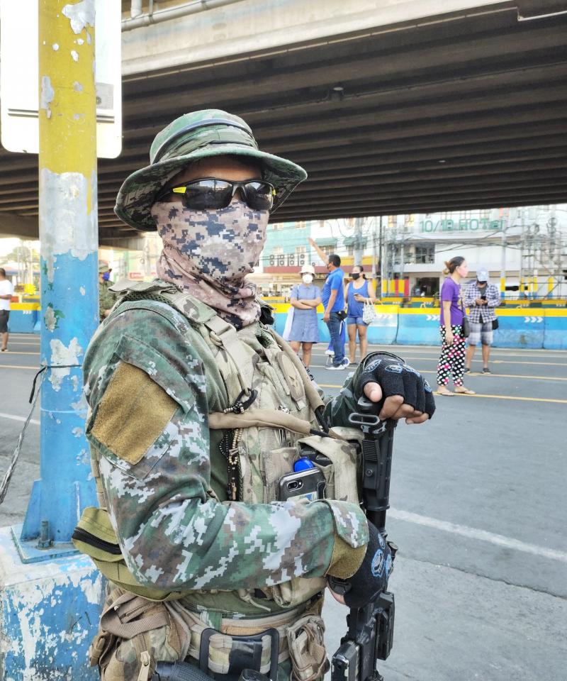 A soldier stands in a street with a gun, wearing a face mask.