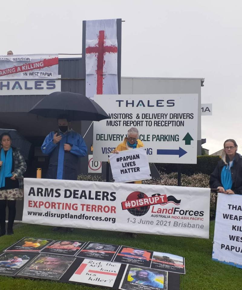 A number of people hold banners in front of a Thales building. One person is on the roof holding a banner.