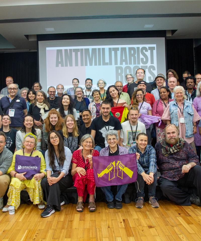 A group photo from Antimilitarist Roots. Around 90 peple gathered on a stage in front of a projected sign with the conference logo