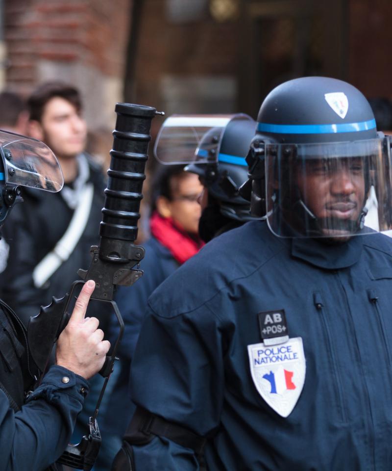Two police officers in the foreground, one armed with a large tear gas launcher