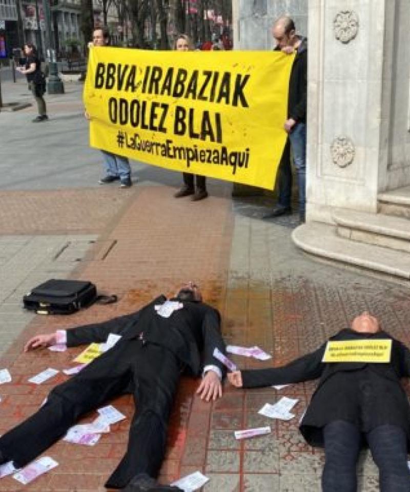 Dos personas vestidas como ejecutivos están tendidas en el suelo, que está manchado con pintura roja. Al fondo, hay personas sosteniendo una pancarta que dice 'La guerra empieza aquí'