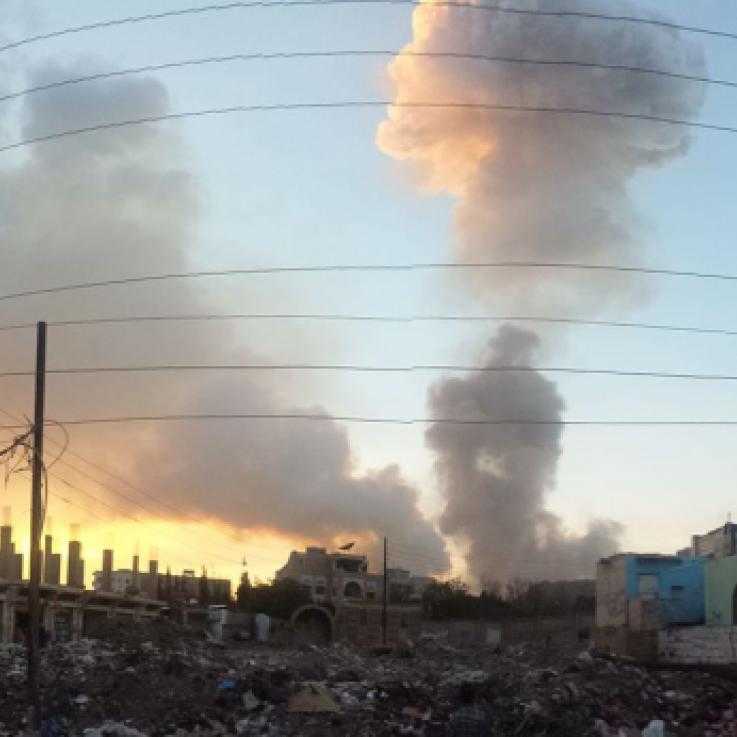 An airstrike in Sana'a, Yemen, in 2015. A cloud of smoke rises from behind buildings. The sun is rising to the left of the picture.