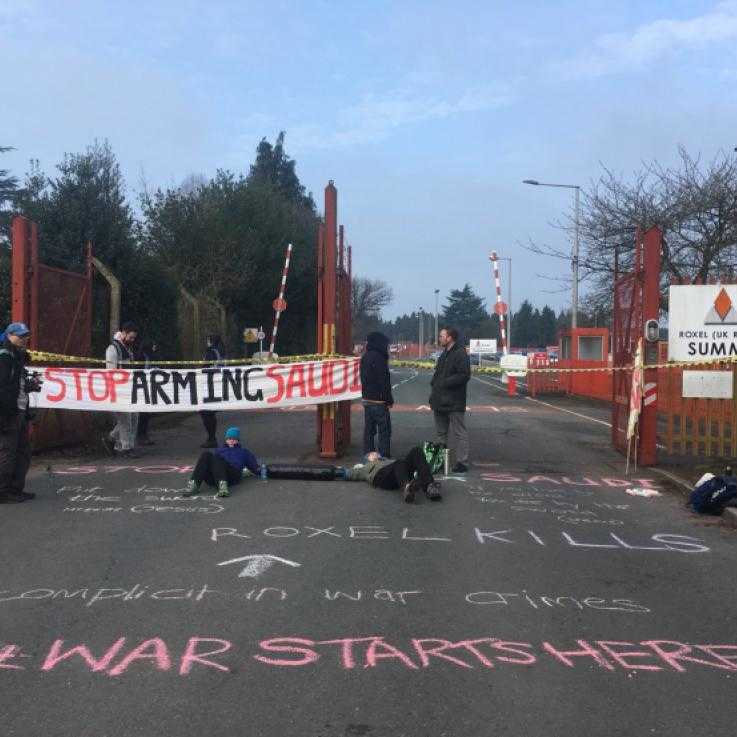 Activists hold a "stop arming saudi" banner across a gate that is blocked by people in a lock-on tube. The floor has been chalked with the words "War Starts Here"
