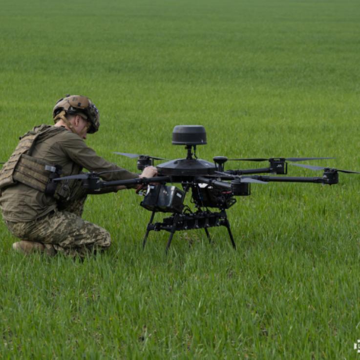 A soldier prepares a drone for flight. He has his back to the camera. They are positioned on grass.