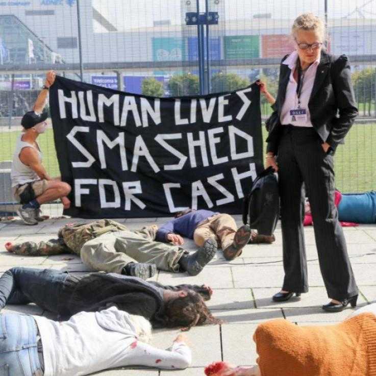 A die-in at DSEI 2017. People lie on the floor, while a woman dressed in business clothing looks down. Behind her activists hold a sign that reads "Human lives smashed for cash"